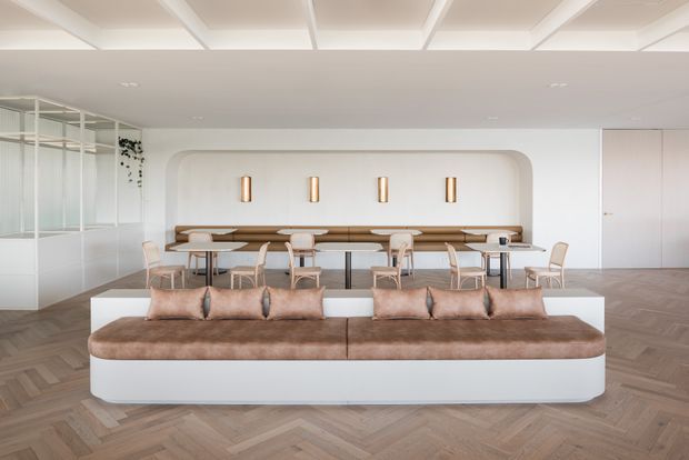 A bright, refined open-plan interior centred on a large curved upholstered banquette or sofa in front of a long dining table. The room features a warm herringbone timber floor, white walls and ceiling, a sculptural recessed wall niche with rounded corners, slim vertical brass-toned wall lights, pale timber and upholstered dining chairs, and a glazed partition at left. The space has a clean contemporary hospitality or residential dining-lounge character.