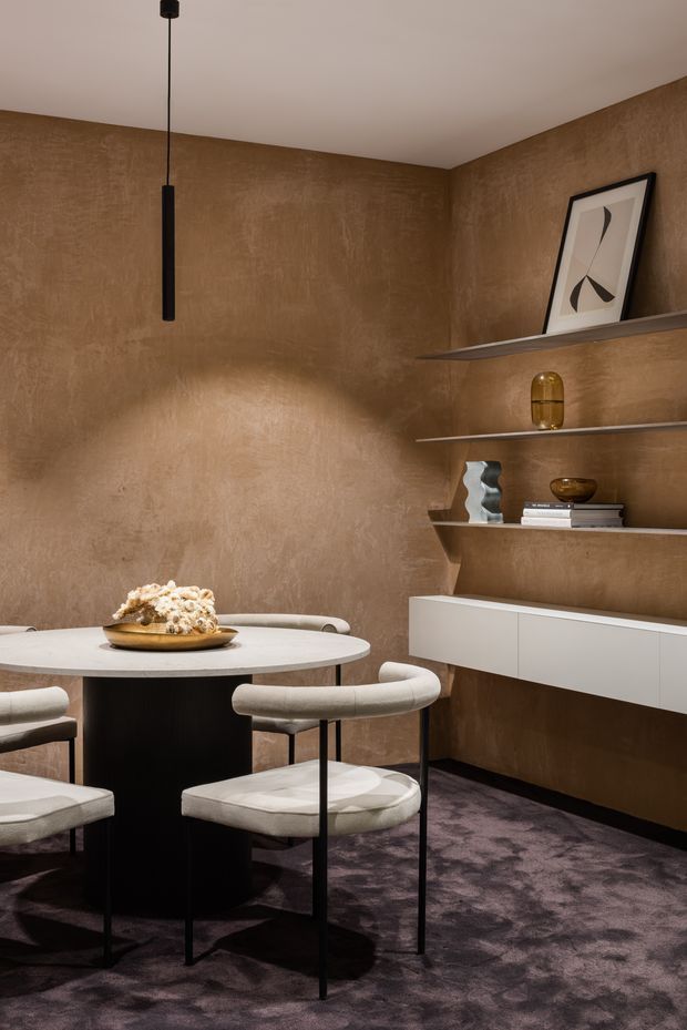 A refined dining area with a round white stone-look table, upholstered curved dining chairs, and a black pedestal base. The room features warm textured brown plaster walls, dark patterned carpet, minimalist black pendant lighting, and a floating white built-in sideboard beneath open display shelves with sculptural decor and artwork. The overall composition is intimate, warm, and contemporary with a luxurious, curated feel.