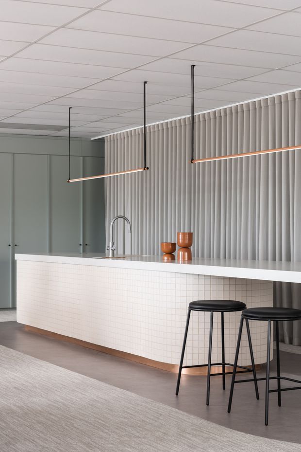 A refined contemporary kitchen interior dominated by a long white island with a subtly gridded tiled front, pale stone-looking benchtop, integrated sink and curved tap. Tall muted grey cabinetry lines the left wall, while a full-height pleated vertical feature wall forms the backdrop. Slim suspended linear pendant lights with warm copper-toned details hang above the island, accompanied by two black bar stools. The palette is restrained in white, grey, black and warm metallic accents, with a large neutral rug in the foreground.