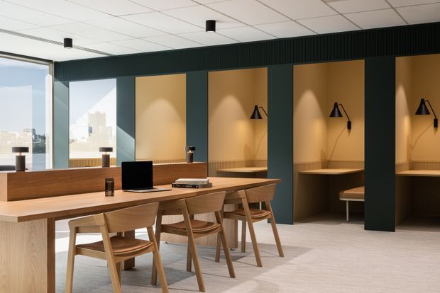 A contemporary open-plan workspace or study area with long light-wood desks, matching wooden chairs, integrated desk lamps, and built-in individual workstations set into a dark charcoal wall. Large windows provide daylight and an urban view, while warm wood panels and focused lighting create a refined, calm office environment.
