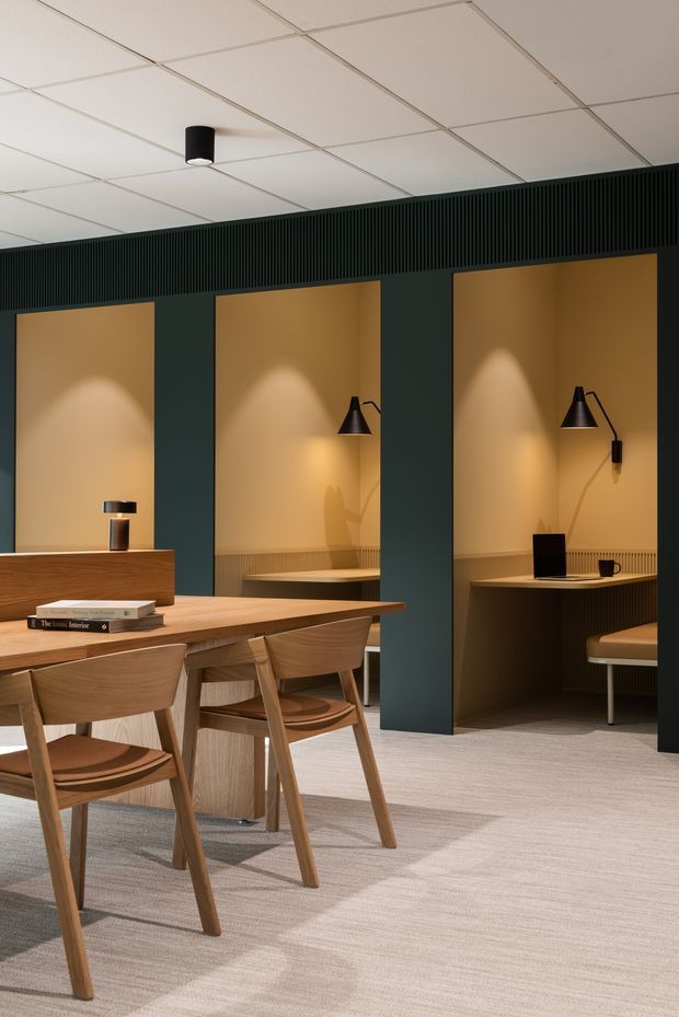 A refined contemporary commercial interior featuring a shared timber dining or meeting table with matching wooden chairs in the foreground. Along the dark green wall are several recessed booth-like workspaces or seating alcoves, each finished in warm tan panels with integrated black task lighting and ribbed detailing. The space has a light carpeted floor, a suspended white grid ceiling, and a warm, subdued palette of timber, tan, deep green, and black.