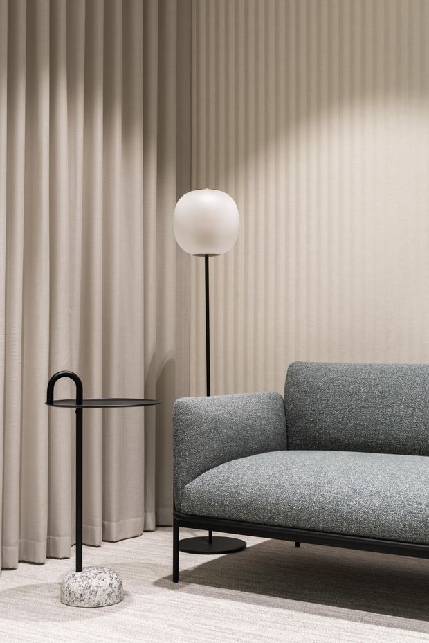 A close-up interior vignette featuring a contemporary grey upholstered sofa with slim black metal legs, a slender black side table, and a tall black floor lamp with a spherical white shade. Full-height pleated beige curtains cover the wall, while a pale textured carpet and darker rug provide layered neutral flooring. The composition is understated, refined, and softly lit.