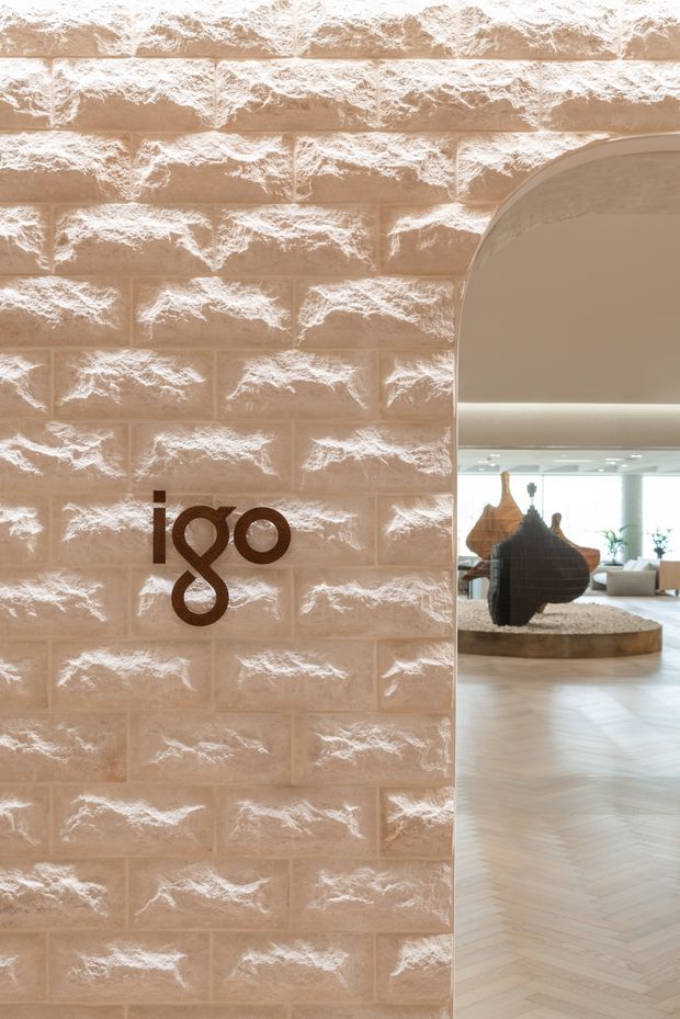 Interior architectural detail showing a warm beige textured masonry feature wall with a rounded arched opening. A dark brown decorative numeral/logo is mounted on the wall. Through the opening, a refined open-plan interior is visible with pale herringbone timber flooring, a circular raised display platform, sculptural decor, and soft recessed lighting.
