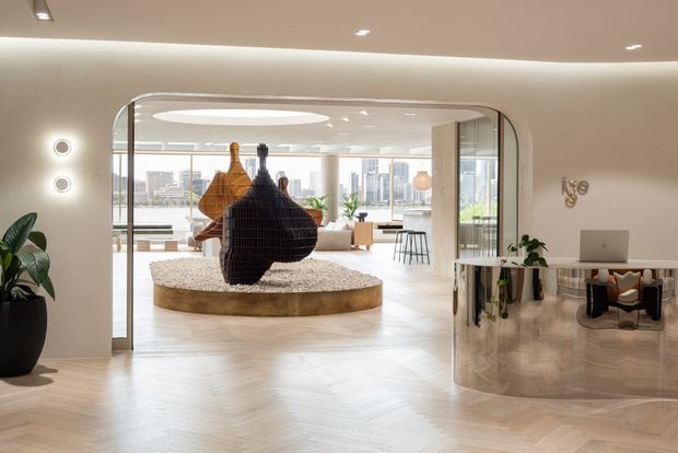 A refined contemporary interior featuring a curved, arched glazed opening leading into a bright open-plan living area. A circular raised platform with large sculptural pendant-like objects forms the centrepiece, while a curved mirrored or reflective built-in desk and workspace occupies the right side. Pale herringbone timber flooring, softly curved walls, recessed lighting, indoor plants, and extensive city-facing glazing create an airy, polished atmosphere.