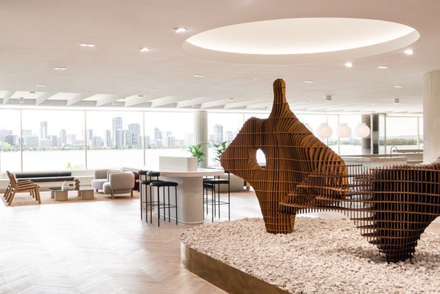 Bright, spacious contemporary commercial interior with floor-to-ceiling glazing overlooking a city skyline and water. The open-plan space features a central dining or meeting counter with stools, lounge seating, pale timber herringbone flooring, recessed lighting, and a large sculptural timber screen or installation set within a raised bed of white stones.