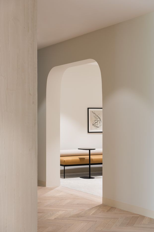 A softly lit minimalist interior viewed through a rounded arched opening. The foreground features a pale timber herringbone floor and a vertically grooved light-toned wall panel. Beyond the opening is a refined sitting area with a long upholstered bench in warm ochre and cream tones, a slender black side table, a pale rug, and framed abstract artwork. The architecture is characterised by curved plasterwork, muted neutral walls, clean lines, and restrained detailing.