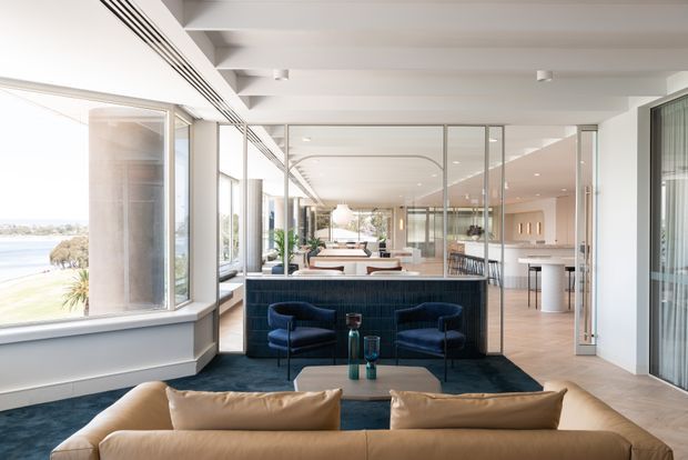 A bright, spacious contemporary open-plan living area overlooking a coastal landscape. The foreground features a tan sofa, while a dark blue tiled or ribbed bar partition separates the seating area from a dining and kitchen zone. Full-height glazing, pale timber flooring, white structural beams, glass partitions, blue armchairs, and layered pendant and recessed lighting create an airy, refined interior.