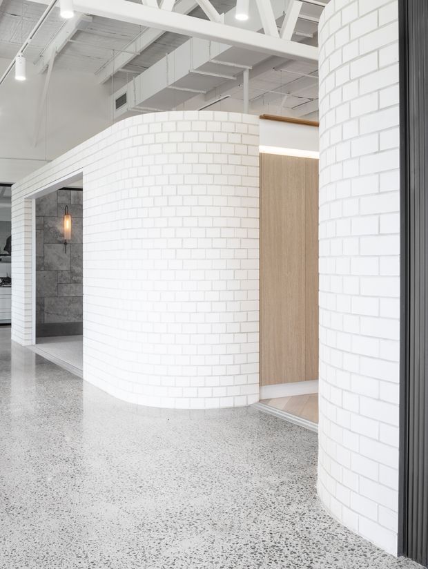 Bright contemporary commercial interior featuring curved white brick partition walls, polished terrazzo flooring, exposed white structural beams and services, and warm timber doors. A tiled service or bathroom area is visible through an opening on the left.