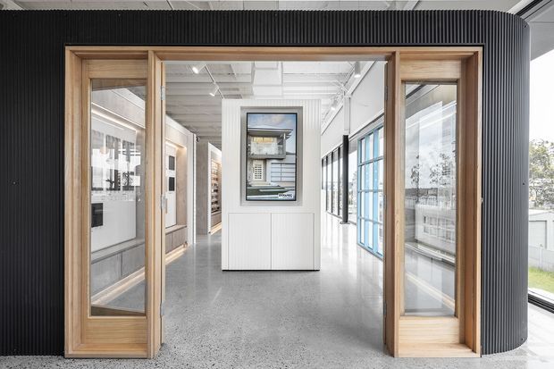 Bright contemporary commercial interior viewed through a timber-framed glazed entry, with a curved black ribbed feature wall, polished concrete or terrazzo-like flooring, exposed white services and ceiling structure, and extensive internal glazing. The space beyond appears to be an office or showroom circulation area with display joinery and a central built-in cabinet.