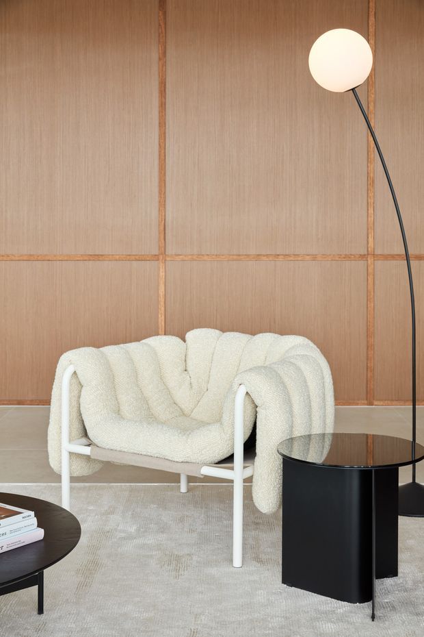 A refined contemporary living room vignette featuring a sculptural cream boucle armchair with a white tubular frame, set against a warm timber-panelled wall with grid-like battens. A slender black arched floor lamp, glossy black cylindrical side table, round dark coffee table, and pale textured rug complete the calm, tonal interior.
