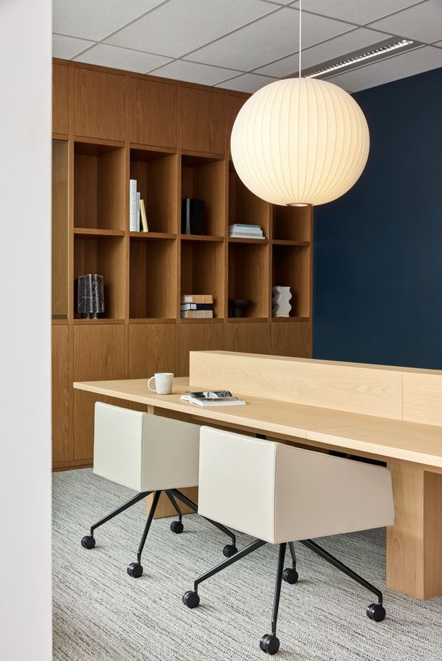 A refined office or study with a long pale timber built-in desk and rear credenza, paired with two white upholstered swivel task chairs on black five-star bases. A full-height dark timber shelving and cabinetry wall provides open display niches and concealed storage. The room features a deep navy accent wall, grey carpet, a suspended spherical paper-style pendant light, and a suspended grid ceiling with linear lighting.