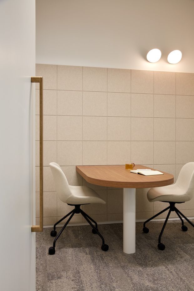 A compact, softly lit office or meeting nook with a rounded wood-grain table, two cream upholstered swivel chairs, and a central white pedestal leg. The back wall is clad in large-format beige textured tiles with visible grout lines, while the floor is covered in grey patterned carpet. A partial glass or reflective partition with a timber-toned frame appears at left, and two round wall or ceiling lights provide warm illumination.