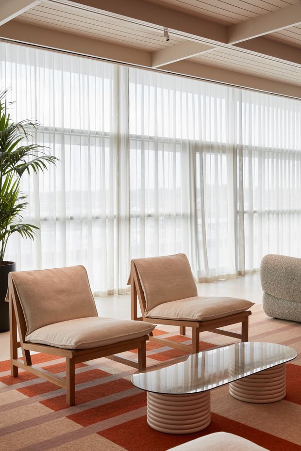 Bright, airy living room with floor-to-ceiling sheer curtains, a glazed wall, exposed white-painted ceiling beams and timber-lined ceiling panels. Two tan upholstered lounge chairs and a rounded grey sofa surround two oval glass coffee tables with ribbed pedestal bases. A geometric terracotta, beige and cream rug adds warmth, while a tall indoor plant softens the corner.