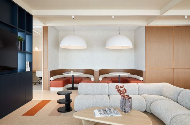 A refined contemporary living room with a large curved light-grey sectional sofa in the foreground, a pale rounded coffee table, and a built-in banquette seating area at the rear. The room features dark navy built-in media and shelving on the left, warm timber cabinetry on the right, textured pale walls, a light timber ceiling, and two oversized pendant lights above the rear seating. Subtle terracotta and muted pink accents add warmth to the neutral palette.