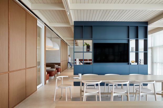 Bright contemporary open-plan dining and living area with a long light-wood dining table and white chairs in the foreground. A deep blue built-in media wall with shelving and television forms the main feature, while warm timber wall panelling, glass partitions, white linear ceiling boards, exposed beams, and full-height sheer glazing create a refined, airy interior.