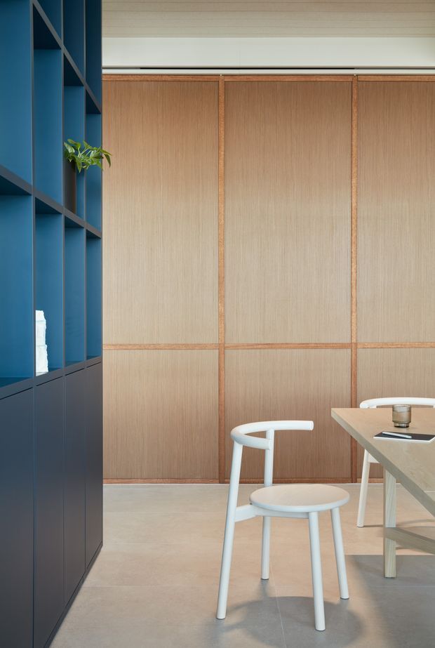 A refined dining area with a pale timber-panelled feature wall, integrated dark navy-blue shelving and cabinetry, and a light stone or timber dining table with white chairs. The space has a restrained contemporary aesthetic, clean vertical and horizontal lines, and a soft neutral palette accented by deep blue joinery and a small indoor plant.