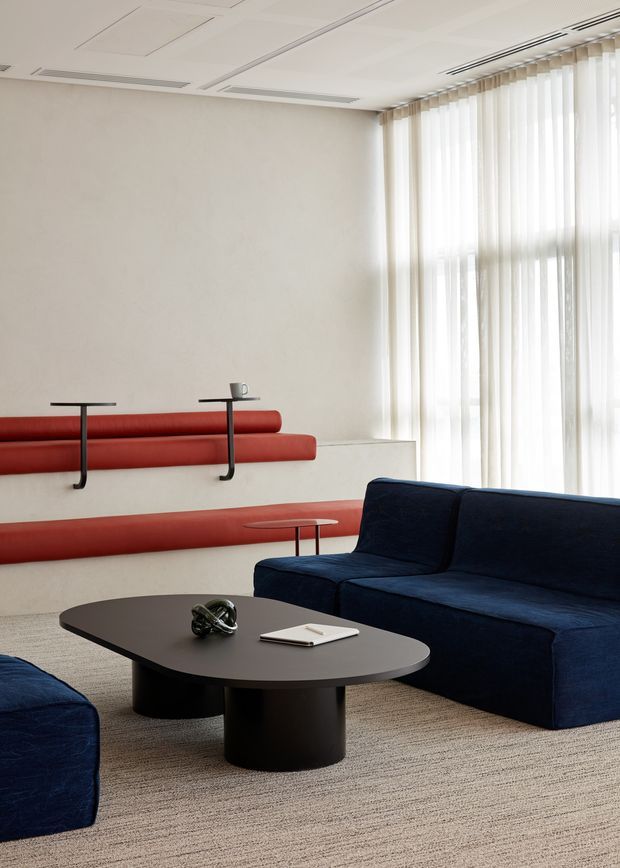 A refined contemporary lounge with deep navy upholstered sofas arranged around a low black oval coffee table. A sculptural red built-in bench runs along the rear wall with slim black supports and small integrated tables. Full-height sheer vertical blinds provide soft daylight, while the pale textured wall, light carpet, and recessed linear ceiling services create a calm, minimalist commercial-style interior.