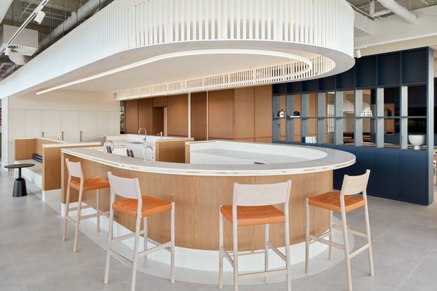 Bright contemporary commercial-style kitchen or hospitality space dominated by a large curved timber island with a pale solid-surface benchtop and integrated seating. The space features white vertical slatted feature detailing and a suspended oval canopy, dark blue built-in cabinetry and open shelving, warm timber wall panels, pale tiled flooring, and exposed industrial ceiling services.