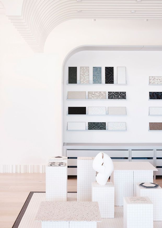 Bright minimalist tile showroom or materials display space with white architectural interiors, curved ribbed ceiling detailing, arched wall niche, and multiple shelves displaying small-format tile samples. The foreground contains tiled display plinths, a counter or table, and a sculptural white object. Pale timber flooring and a light terrazzo-like display surface contribute to the clean, gallery-like setting.