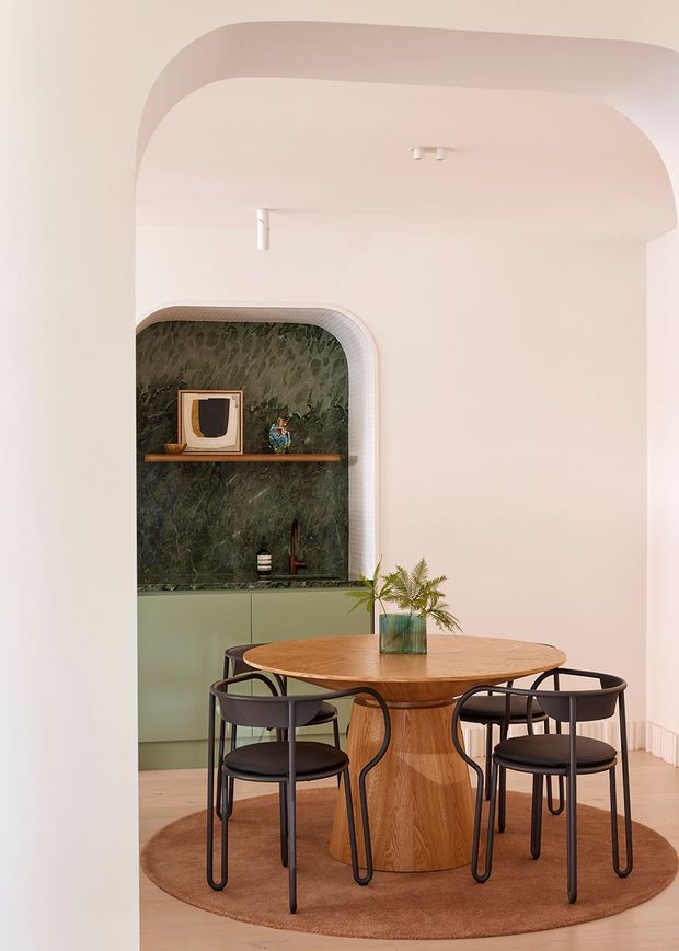 A softly styled dining nook with a round timber dining table and four black-framed chairs, set on a circular brown rug. The room features rounded architectural openings and ceiling edges, pale blush walls, light timber flooring, and a built-in green cabinetry and dark green stone feature niche with a timber shelf and decorative objects.