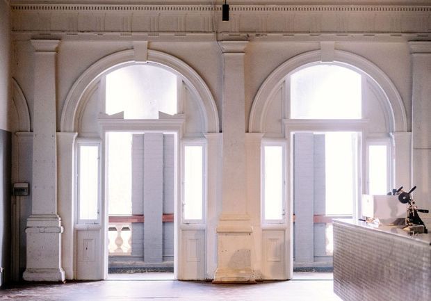 Interior view of a grand, light-filled space with two tall arched glazed doorways framed by ornate white architectural mouldings and pilasters. The openings lead to a balcony or exterior terrace. A tiled built-in counter or reception desk occupies the lower right, with a small lamp or equipment on top. The palette is predominantly white and warm neutral, with strong daylight and traditional architectural detailing.