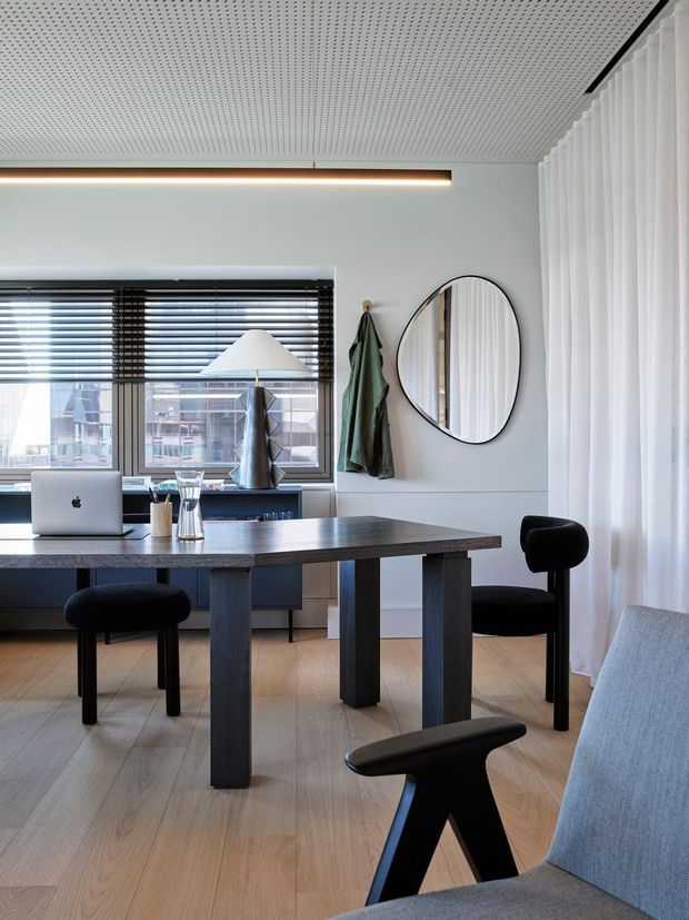 A bright, contemporary home office or work area with a large dark timber desk, laptop, black chairs, and a pale upholstered lounge chair. The room features light timber flooring, white walls, full-height sheer curtains, horizontal window blinds, a round black-framed mirror, and a perforated acoustic ceiling with a linear recessed light.