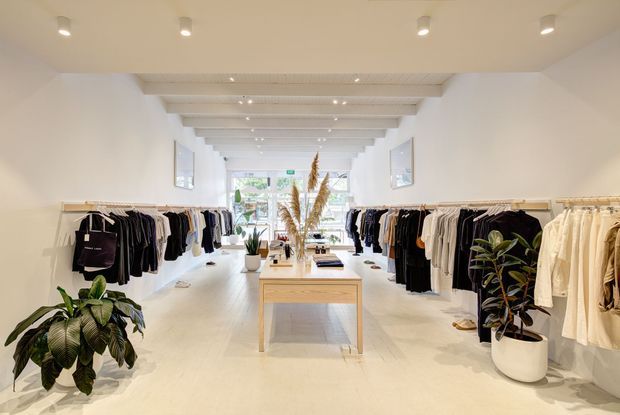 Bright minimalist clothing boutique with white walls and flooring, long timber garment rails displaying clothing along both sides, a central light-wood display table, indoor plants, and a white exposed-beam ceiling with recessed lighting. Large rear glazing brings in natural light and greenery is visible beyond.
