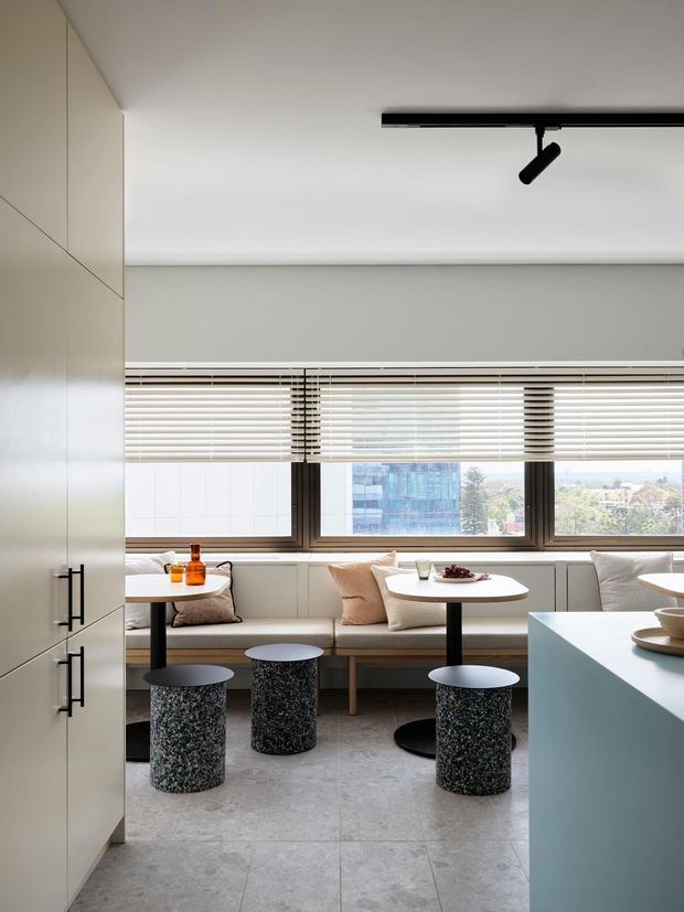 Contemporary open-plan dining and seating area with a built-in window bench, round café tables, textured cylindrical stools, and a pale blue kitchen island or counter in the foreground. Large horizontal windows with timber-toned frames and venetian blinds overlook an urban setting. Minimal white cabinetry and a black ceiling track light complete the clean, modern interior.
