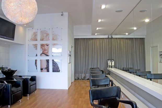 A contemporary hair salon interior with a row of black leather styling chairs facing a long mirrored workstation and counter. The room features warm timber flooring, white walls and ceiling, recessed lighting, full-height grey curtains, and a large spherical pendant light. A television and decorative wall artwork are visible near the seating area.