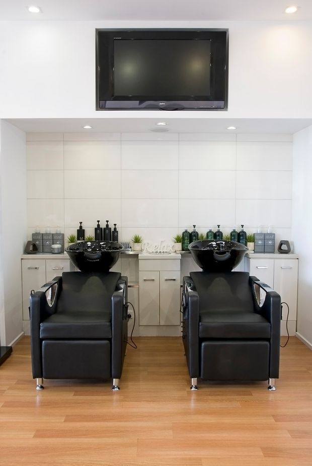 Bright, clean hair salon wash area with two black shampoo chairs and integrated basins facing a long wall of white built-in cabinetry. A wall-mounted television sits above, with salon products and small decorative plants arranged on the counter. The room has recessed lighting, pale tiled wall panels, and warm timber flooring.