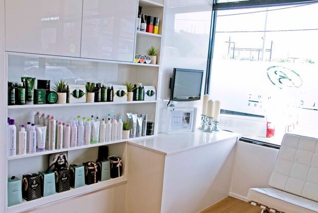 Bright, compact commercial beauty salon reception or retail area with white built-in display shelving stocked with hair and beauty products, a white reception counter, small monitor, decorative plants, and a white upholstered chair. Large front glazing provides abundant daylight, while light timber flooring and glossy white cabinetry create a clean, contemporary fit-out.
