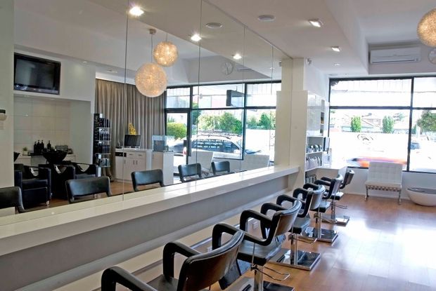 Bright contemporary hair salon interior with a long mirrored styling wall, multiple black salon chairs and stations, pale cabinetry, large street-facing windows, warm timber-look flooring, and spherical pendant lights.