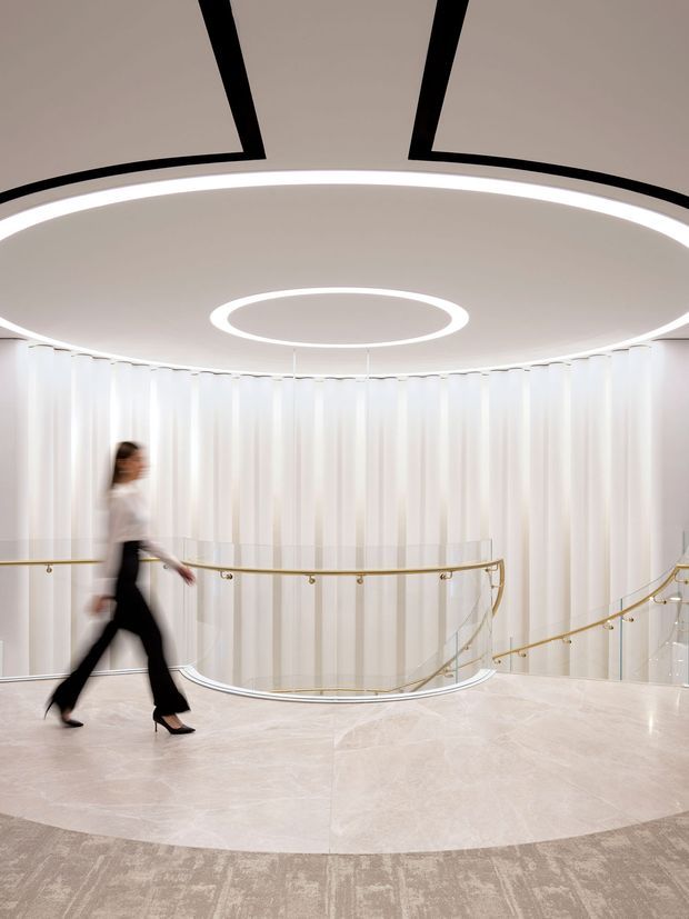 A highly refined curved landing or circulation space overlooking a sweeping staircase. The interior features a luminous sculptural ceiling with large elliptical recessed light rings, sheer white vertical curtains or wall panels, a pale polished stone floor, and a curved glass balustrade with slim brass-gold handrails. A blurred person walking through the space conveys its scale and movement.