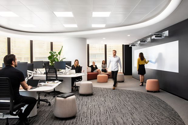 An open-plan modern office with shared workstations, computer monitors, rolling office chairs, and a large presentation wall. The room includes a casual collaboration area with upholstered ottomans and seating, extensive windows with timber blinds, recessed ceiling lighting, and a curved illuminated ceiling detail. Several people are using the workspace and presentation area.
