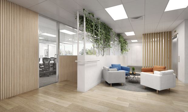 Bright contemporary office reception and lounge area with pale timber flooring, vertical timber slat wall panels, glazed meeting or workstation area, white partition walls, suspended greenery, and a seating arrangement of white, blue, and tan lounge chairs around a small coffee table. The space has a suspended acoustic tile ceiling with recessed fluorescent lighting and a clean professional fit-out.