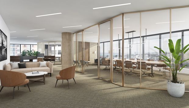 A bright contemporary open-plan commercial office interior with a carpeted lounge reception area in the foreground and a glass-partitioned meeting or workroom beyond. The space features floor-to-ceiling glazing, slim timber-toned frames, exposed concrete columns, linear recessed ceiling lights, desks and chairs, a neutral lounge setting, and a large indoor plant. Urban buildings are visible through the windows.