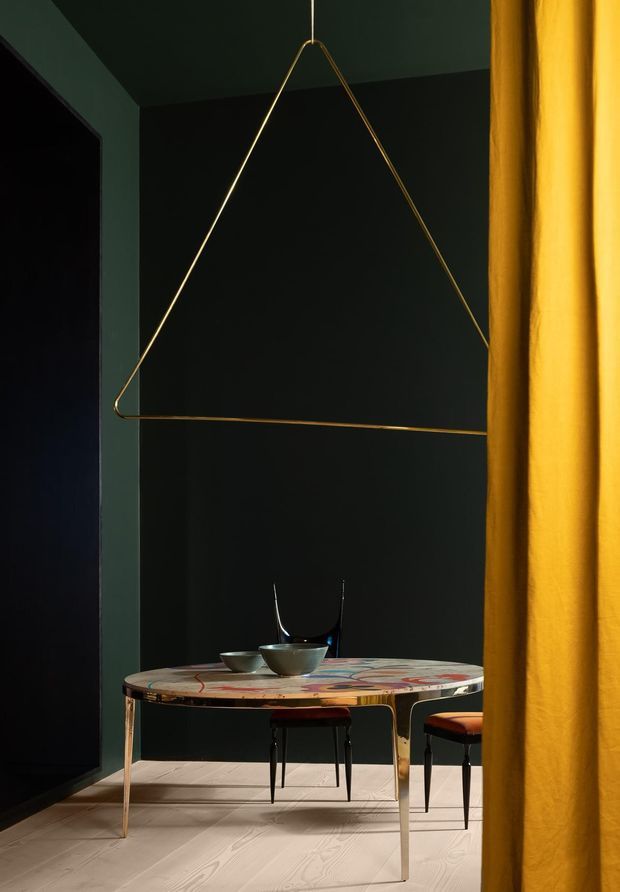 A moody, dramatically styled dining area viewed past a mustard-yellow curtain. The space has deep green-black walls and ceiling, pale timber flooring, a round or oval dining table with a colourful patterned top and metallic legs, dark dining chairs, and a striking suspended geometric brass pendant frame. The composition is intimate, high-contrast, and richly coloured.