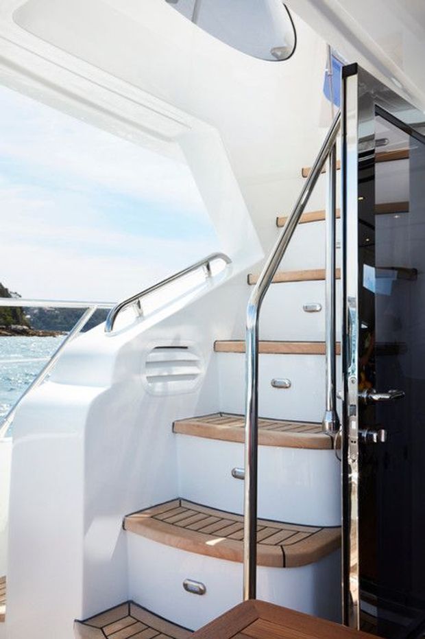 A bright, polished yacht interior showing a compact staircase with white molded walls, teak-topped treads, stainless-steel handrails, and integrated step storage. A glossy dark doorway opens into the cabin, while blue water and coastline are visible outside.