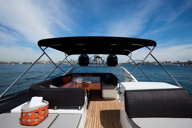 A covered motorboat cockpit viewed from the stern, featuring a teak-style deck, wraparound dark upholstered seating, a central dining table, and a bright waterfront city skyline in the background. The image is a clear outdoor marine leisure setting under a blue sky.