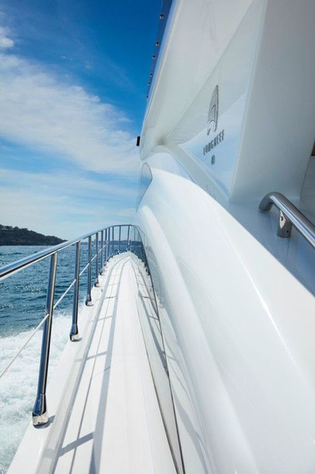 A bright exterior view along the narrow side deck and stainless-steel railing of a large white luxury yacht moving through blue coastal water. The streamlined white hull and superstructure dominate the frame, with a partial yacht name/logo visible on the side and a wooded shoreline in the distance.