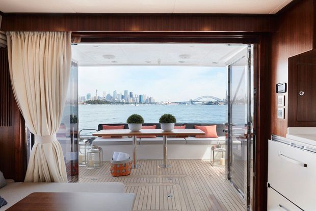 Interior view from a compact, richly timber-lined cabin or boat-like space opening onto a bright covered deck. The deck has built-in bench seating, a narrow table, potted plants, and a pale slatted floor, with a waterfront city skyline and large arch bridge visible beyond. Cream curtains frame the wide glazed opening, while white built-in cabinetry and controls line the right wall.