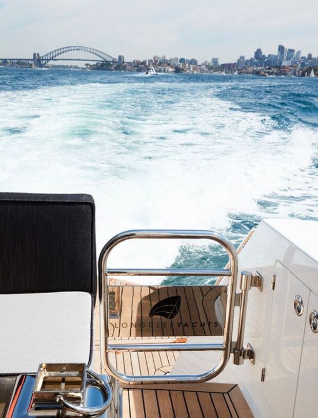 A view from the stern of a luxury motor yacht underway, showing wake-filled blue water, teak decking, polished stainless-steel railings, white marine cabinetry, and upholstered seating. Sydney Harbour Bridge and the city skyline are visible in the distance.