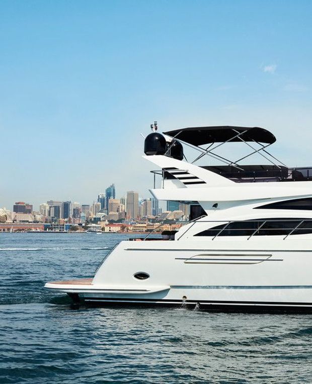 A close-up exterior view of a large white luxury motor yacht cruising on blue water, with a modern city skyline visible in the background. The yacht features multiple decks, dark tinted glazing, sleek streamlined forms, and an upper canopy.