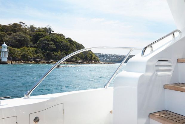 Bright view from the aft deck of a white motorboat, with stainless-steel rails, built-in steps, and timber-trimmed treads. The boat is cruising or moored on clear blue coastal water beside a wooded shoreline, with a lighthouse visible at the left.
