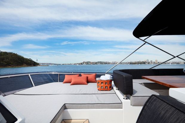 A modern yacht deck with a spacious light-grey upholstered sun lounge, orange cushions, dark woven seating, and a wood-topped dining table beneath a black canopy. The boat is positioned on calm harbour water with a distant urban shoreline visible beyond.