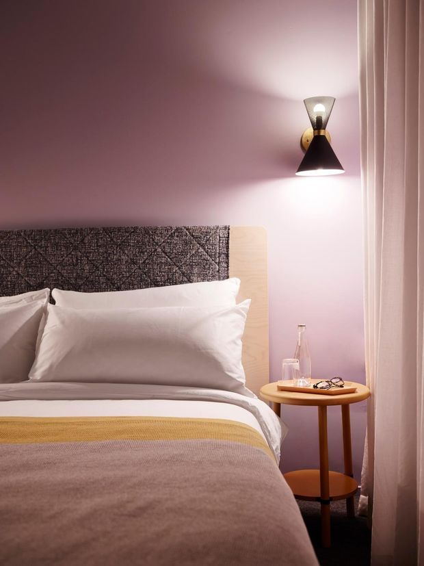 A softly lit bedroom with a bed dressed in white linens and layered mauve, beige, and mustard bedding. The room has dusty pink walls, a dark textured upholstered headboard, pale timber bed detailing, a warm wood-and-brass-toned round side table, a wall-mounted sconce, and sheer floor-length curtains.