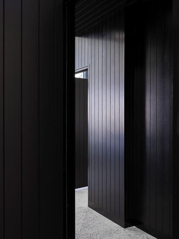 A dark, narrow architectural passage or entry area lined with vertically grooved black wall panelling. A partially open black door reveals a brighter adjoining space, with a small horizontal window and a pale speckled floor providing contrast. The image has a minimalist, moody, highly graphic character.