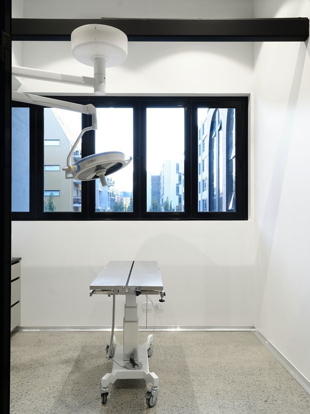Bright, clinical treatment or operating room with white walls, black-framed windows, a mobile examination or operating table, and an overhead surgical light. The room has a polished speckled hard floor and views of surrounding urban buildings through the windows.