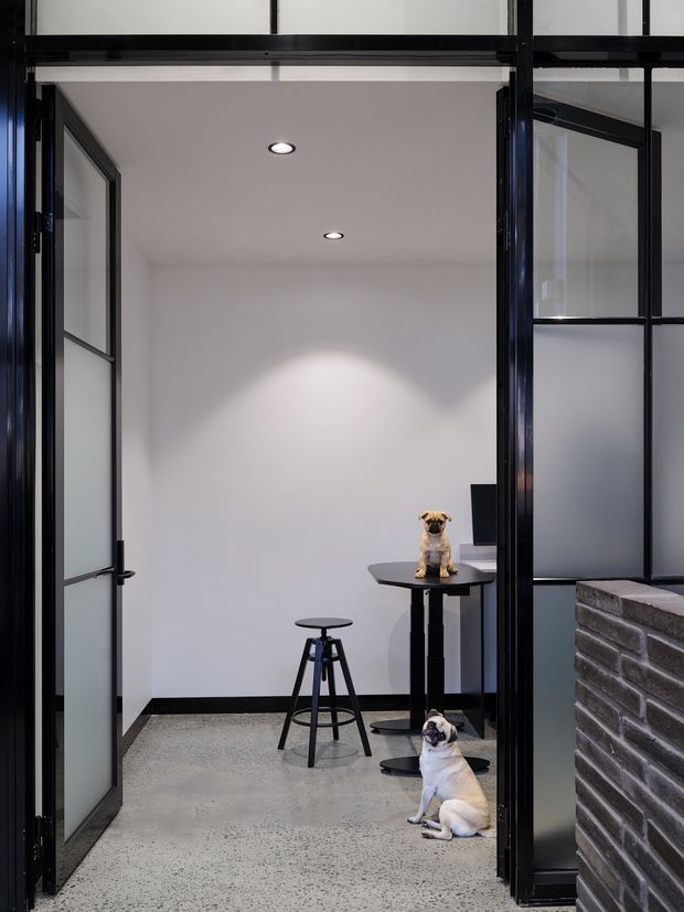 A compact, minimalist interior nook viewed through open black-framed glazed doors. The space has white walls, recessed ceiling lights, a polished speckled concrete floor, a small round black table with a stool, and a dark screen or monitor. A black brick-clad half-height wall is visible in the foreground, with two dogs in the room.
