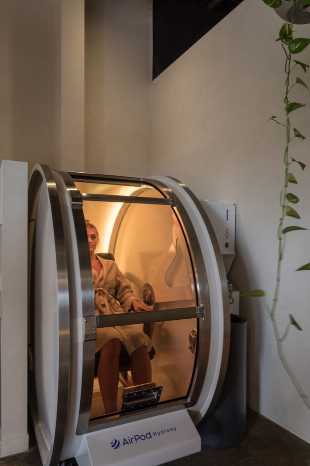 A compact commercial wellness treatment space featuring a person seated inside an AirPod Hydroxy capsule. The capsule has a curved white body, brushed metal trim, transparent door, warm amber interior lighting, and a wall-mounted control panel. A trailing green plant hangs beside it, with neutral plaster walls and dark flooring.